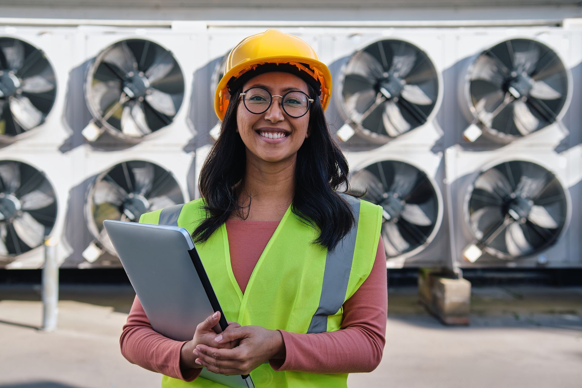 Woman in hard hat and safety vest smiles, holding tablet in front of industrial fans.