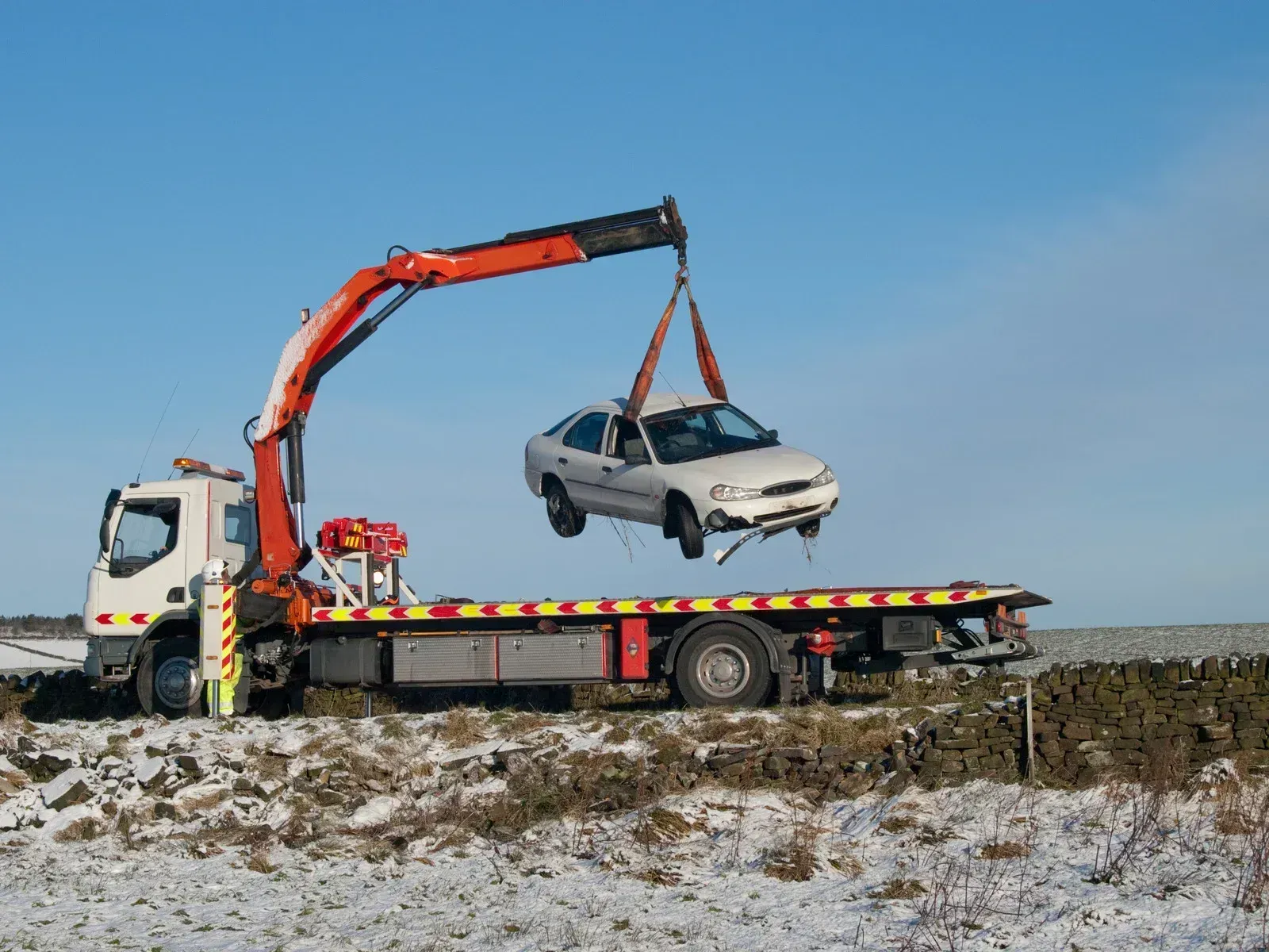 Tow truck lifting a damaged white car in a snowy field on a sunny day.