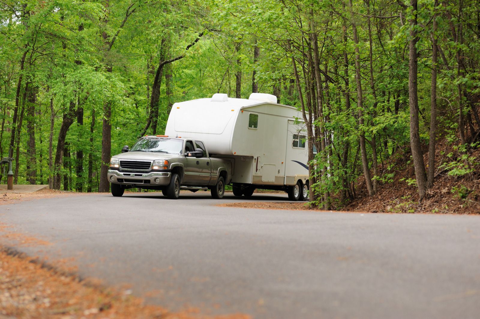 Silver pickup truck towing a white camper on a paved road through a green forest.