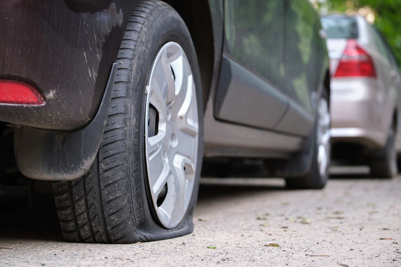 Flat tire on a dark-colored car, parked on a gravel surface next to a tan car.