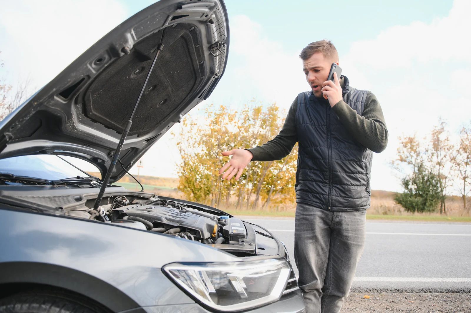 Man on phone beside car with open hood, gesturing in a field.