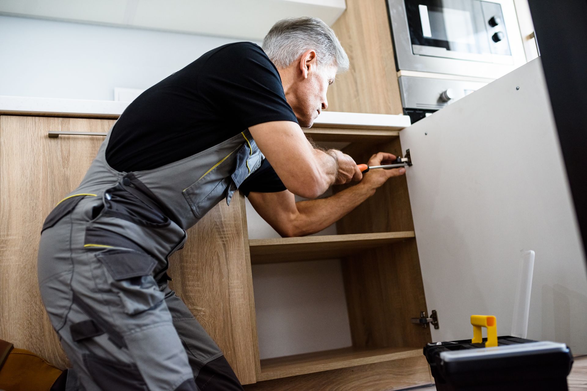 An elderly repairman in a uniform diligently fixing a kitchen cabinet using a screwdriver.