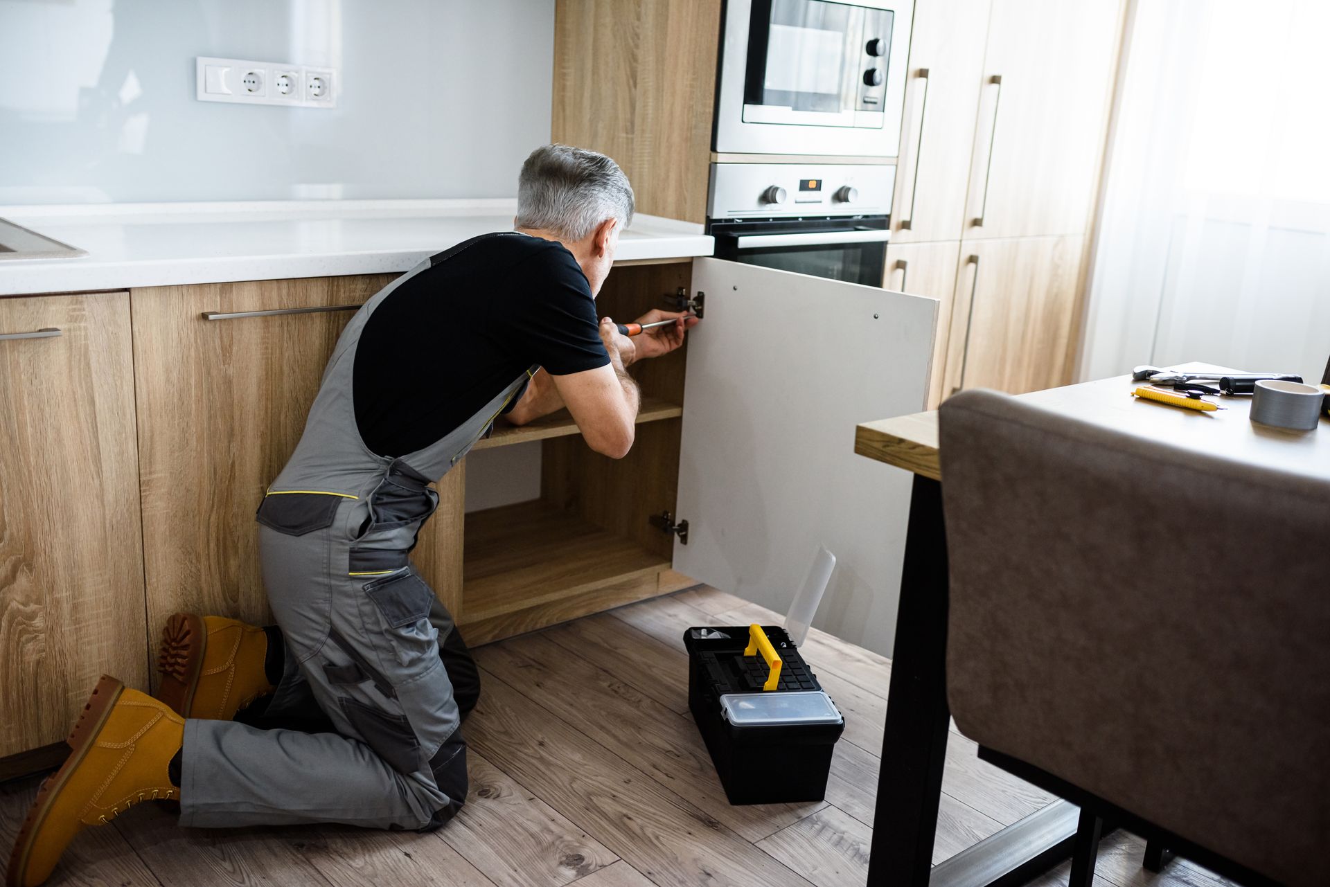 Elderly repairman in uniform skillfully fixing kitchen cabinet with a screwdriver.