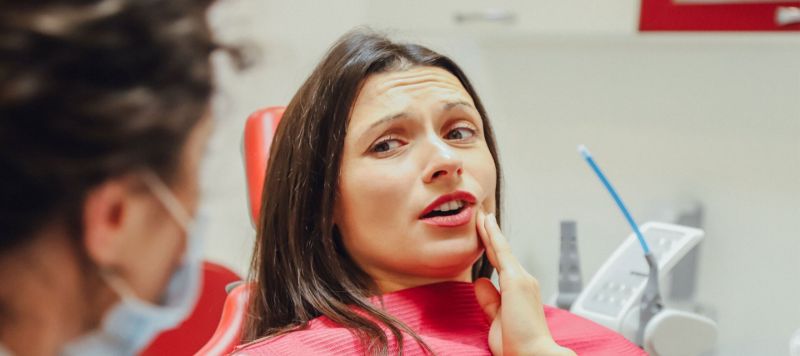 Woman in a dentist chair grimaces, touching jaw. A dentist in a mask is visible in the background.