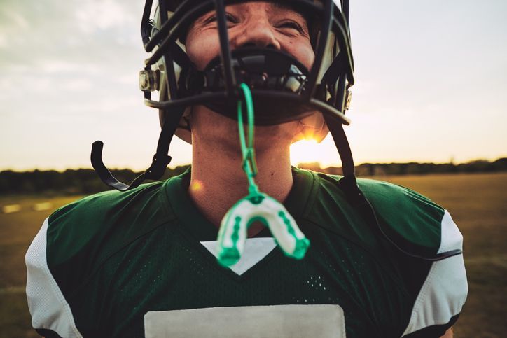 Football player with helmet and mouthguard, smiling on a field at sunset. Green jersey.