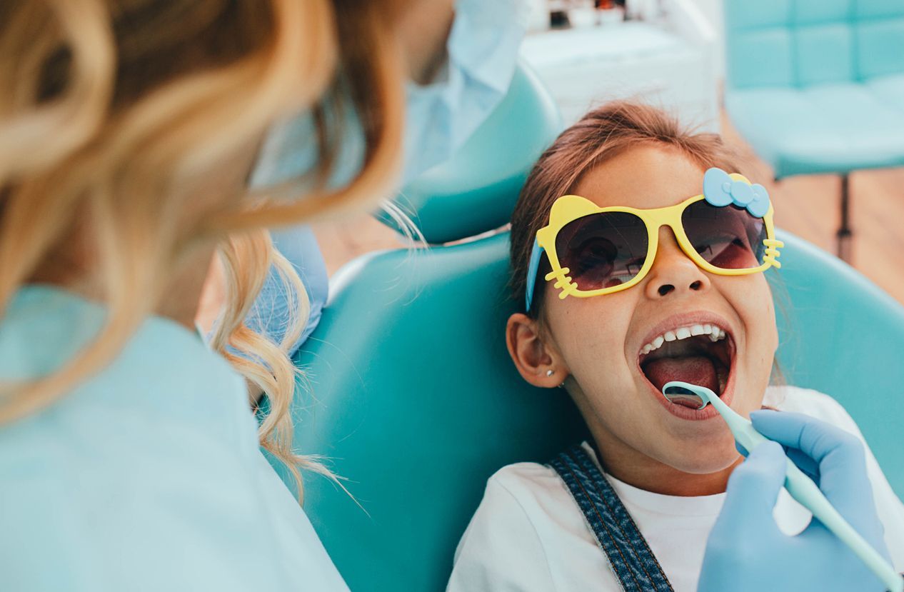 Child with sunglasses at dentist, mouth open, dentist examining teeth.