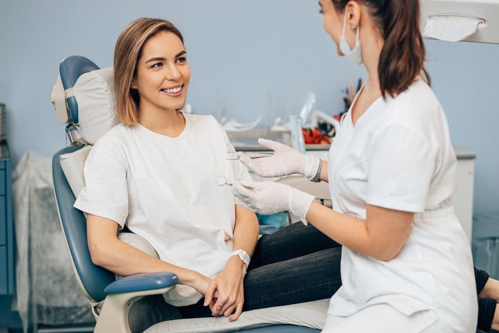 Woman smiling at dentist in dental chair, light-colored office. Dentist gestures, wearing gloves and mask.