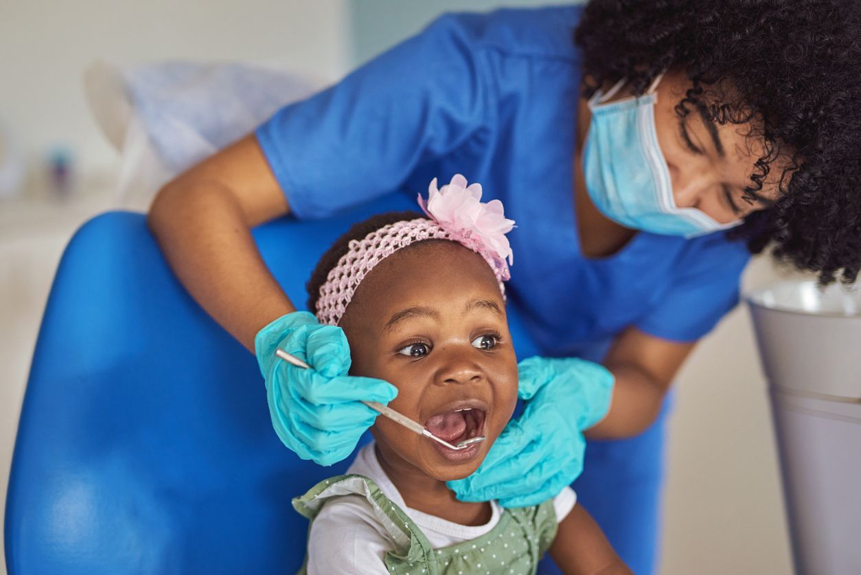 Dentist examining a child's mouth with a dental tool. Both are wearing gloves and masks in a dental office.