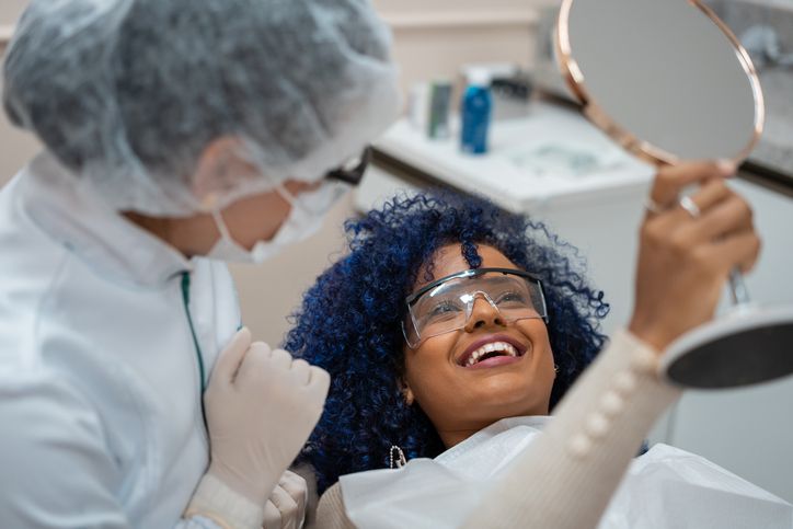 Patient smiling, looking at teeth in mirror after dental work; dentist nearby.