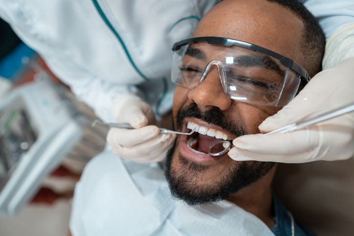 Person at dentist's, mouth open, being examined. Dental tools and hands are visible. The person wears safety glasses.