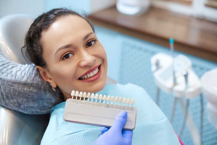 Woman in dental chair smiles, comparing teeth shade with dentist's color chart.