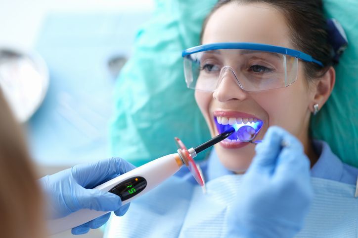 Dentist using blue light on patient's teeth. Patient wearing safety glasses smiles in a dental chair.
