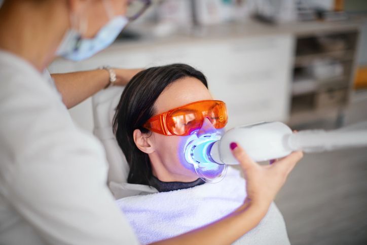 Woman undergoing teeth whitening with blue light. Dentist in mask holds the device.