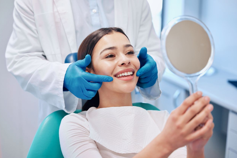 Dentist examining patient's teeth. Patient smiles, holding a mirror. Dental office setting.
