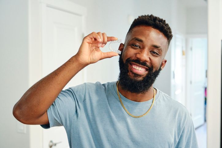 Man smiling, holding up a small, clear disc. Wearing a blue shirt and gold chain, indoors.