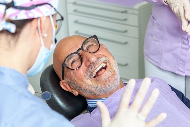 Man in dental chair laughs, dentist in mask and gloves looks on, white and lavender tones.