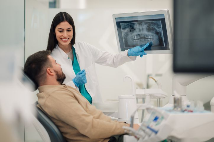 Dentist pointing at X-ray on screen, explaining to patient in dental chair.