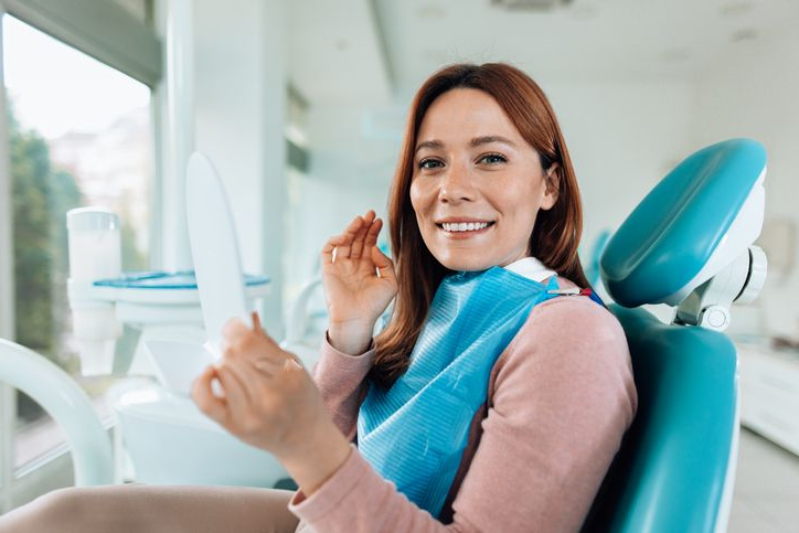 Woman in dental chair smiles while holding a mirror, looking at her teeth.