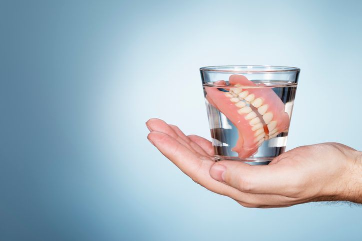 Hand holding a glass of water with dentures submerged in it. Light blue background.
