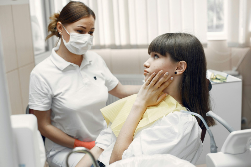 Dentist in mask comforts patient in dental chair, who holds her cheek. Yellow drape, white setting.