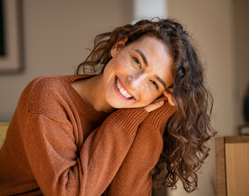 Woman with curly hair smiles, leaning on arms, wearing an orange sweater.