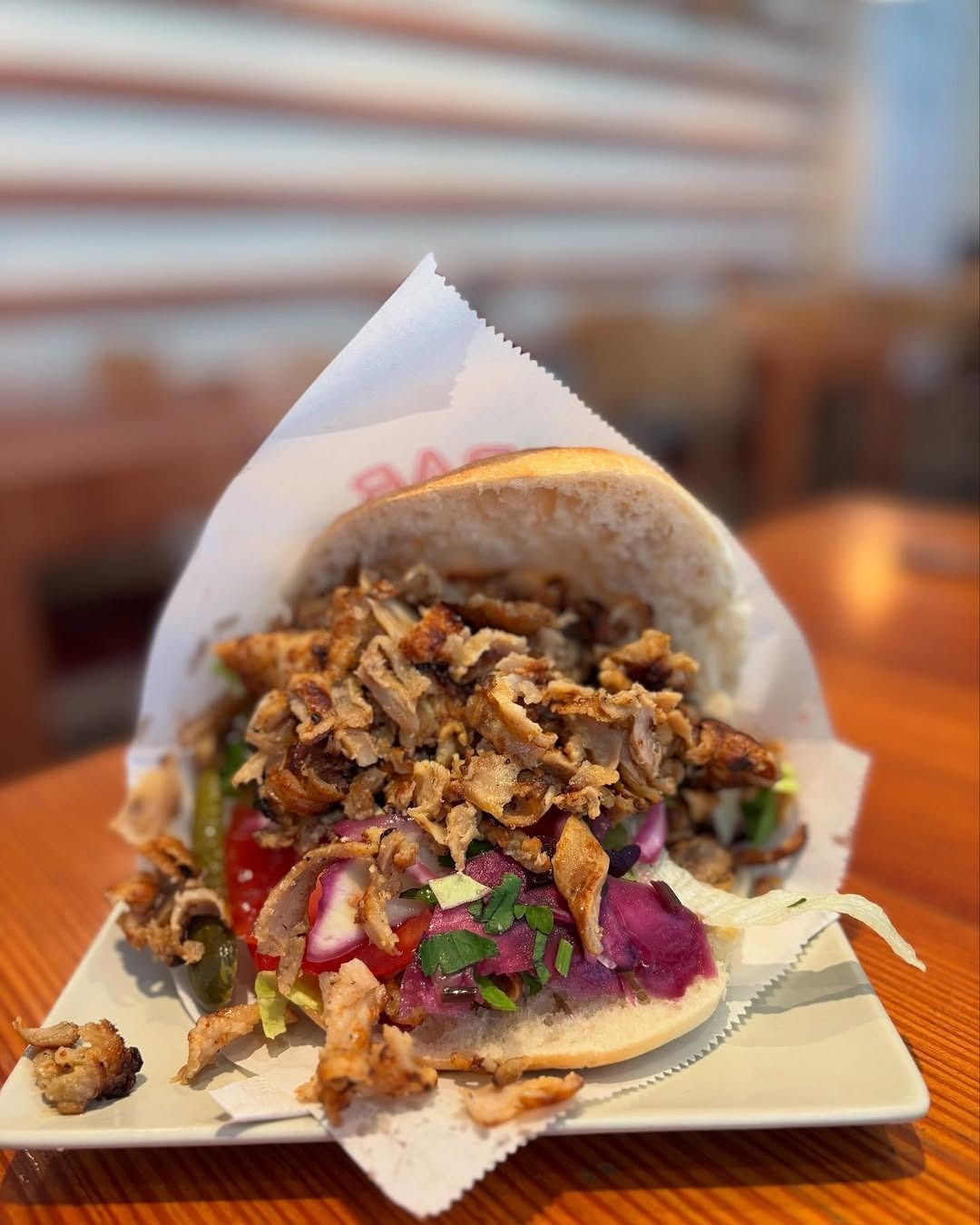 Panino con kebab su un piatto, che fuoriesce da un foglio di carta bianca. Carne, verdure e salsa sono visibili. Tavolo di legno.