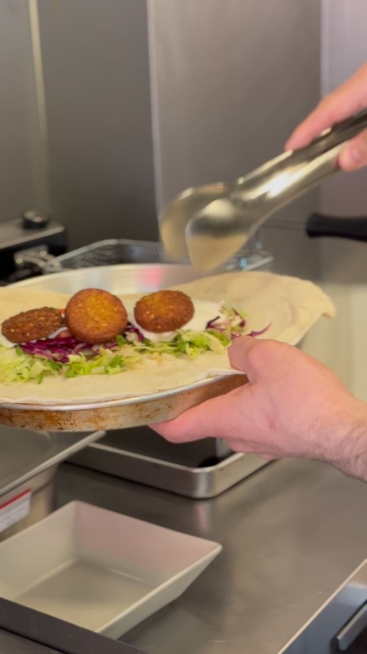 Persona che, con le pinze, dispone le palline di falafel su una piadina in cucina.