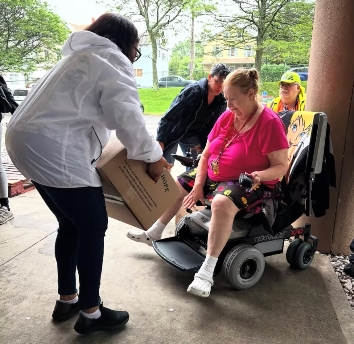 A person hands a cardboard box to someone seated in a motorized wheelchair under an outdoor covered walkway.