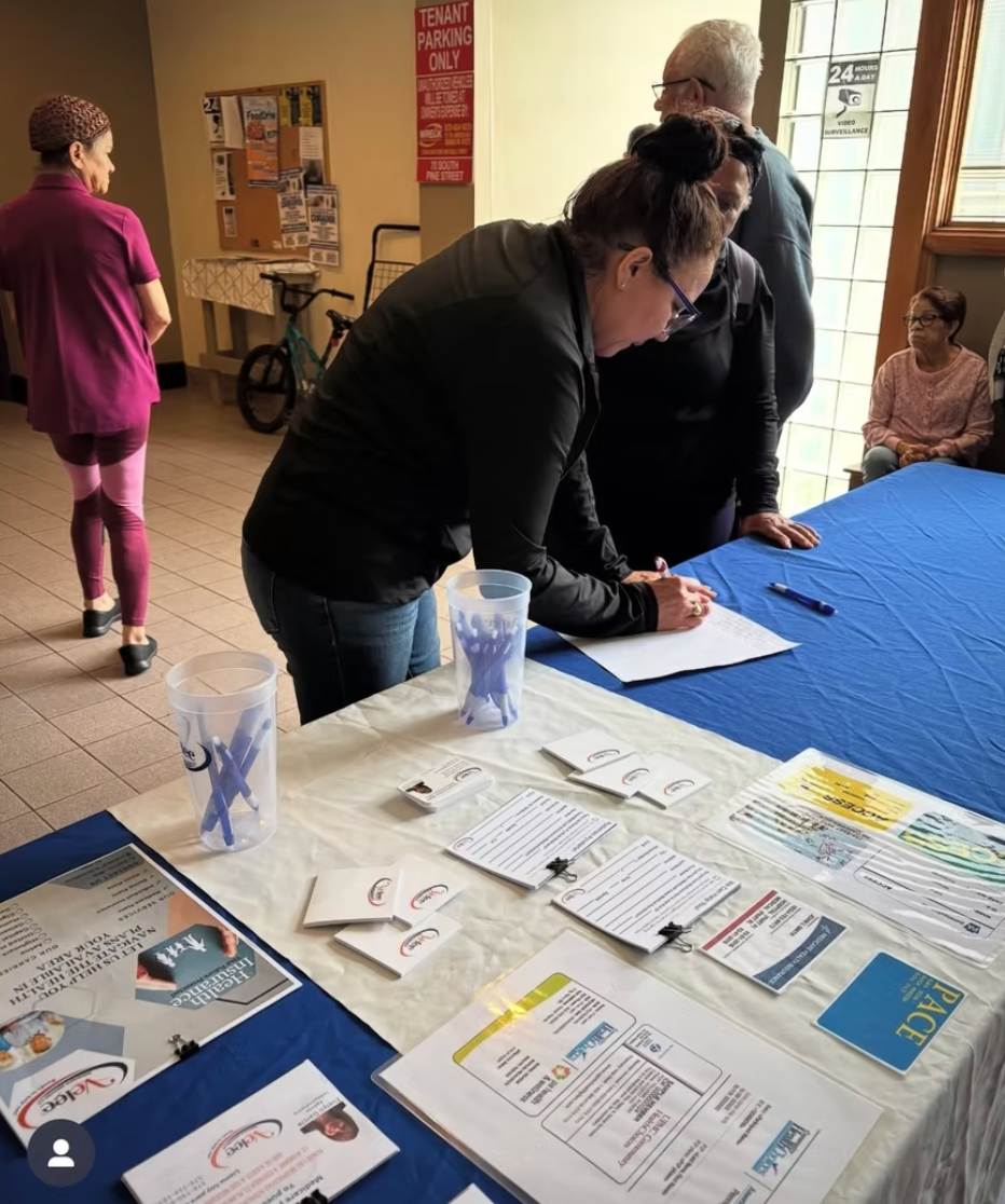 People gather around a table with sign-in sheets and information flyers at an indoor event.