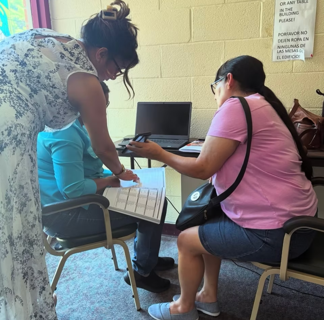 Three people collaborate at a table with a laptop and paperwork in an indoor office or community space.