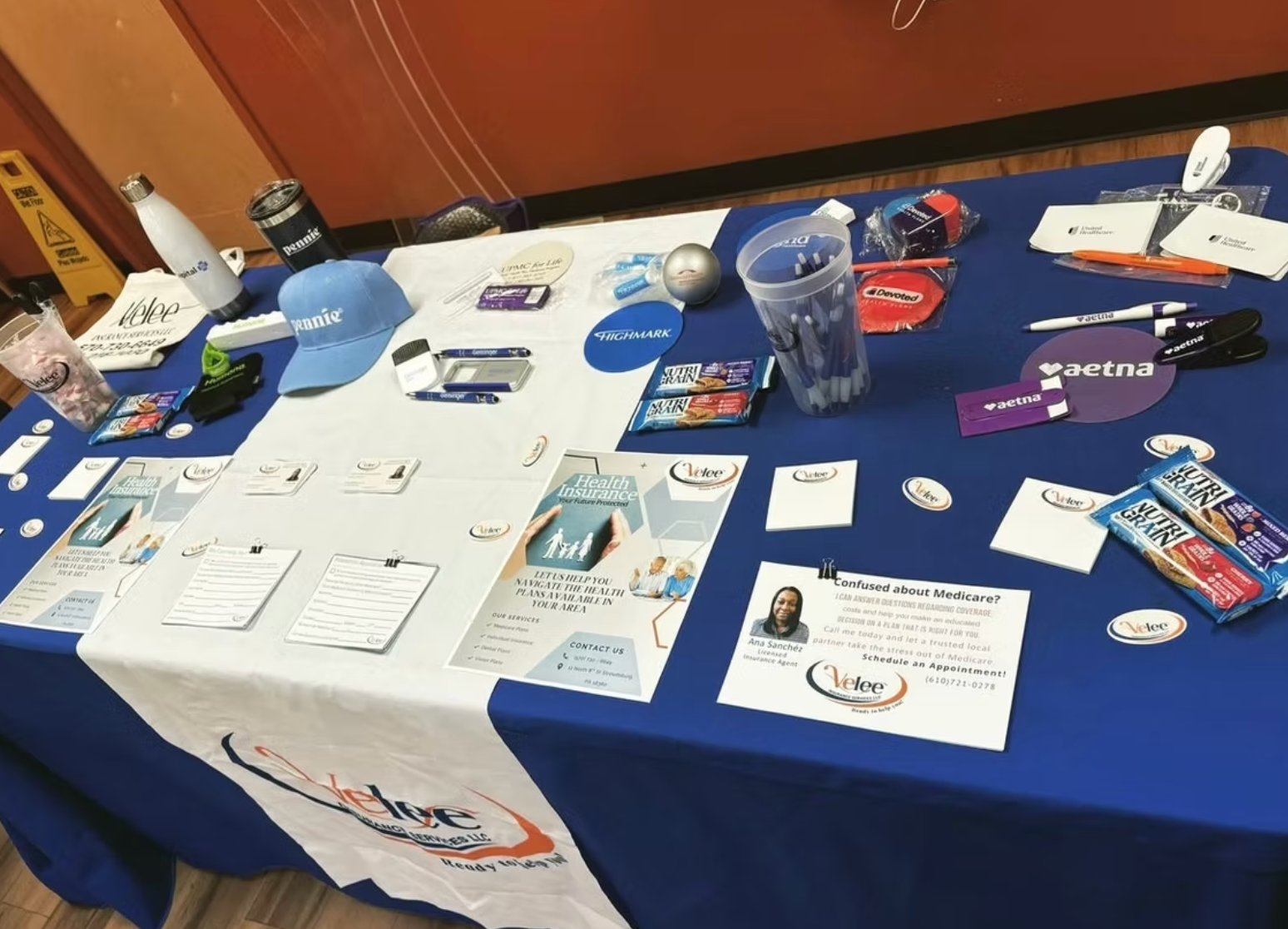 A blue display table at a community event featuring branded pens, brochures, health information, and promotional items.