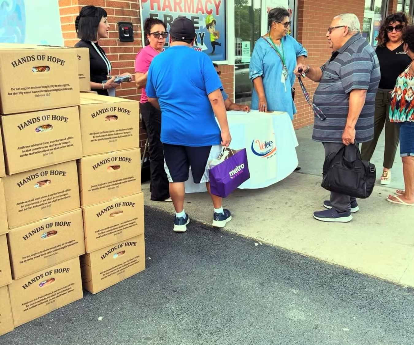 A group of people gathers around a folding table outside a building with stacks of 
