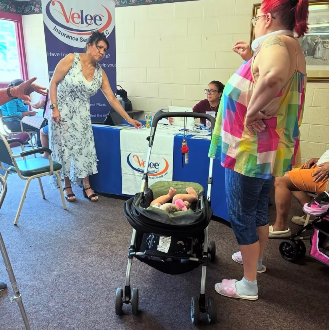 A woman in a floral dress gestures at an information table; another person stands with a baby in a stroller.
