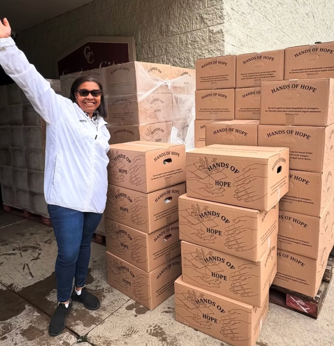 A person with a cheerful expression stands with an arm raised next to stacks of boxes labeled 