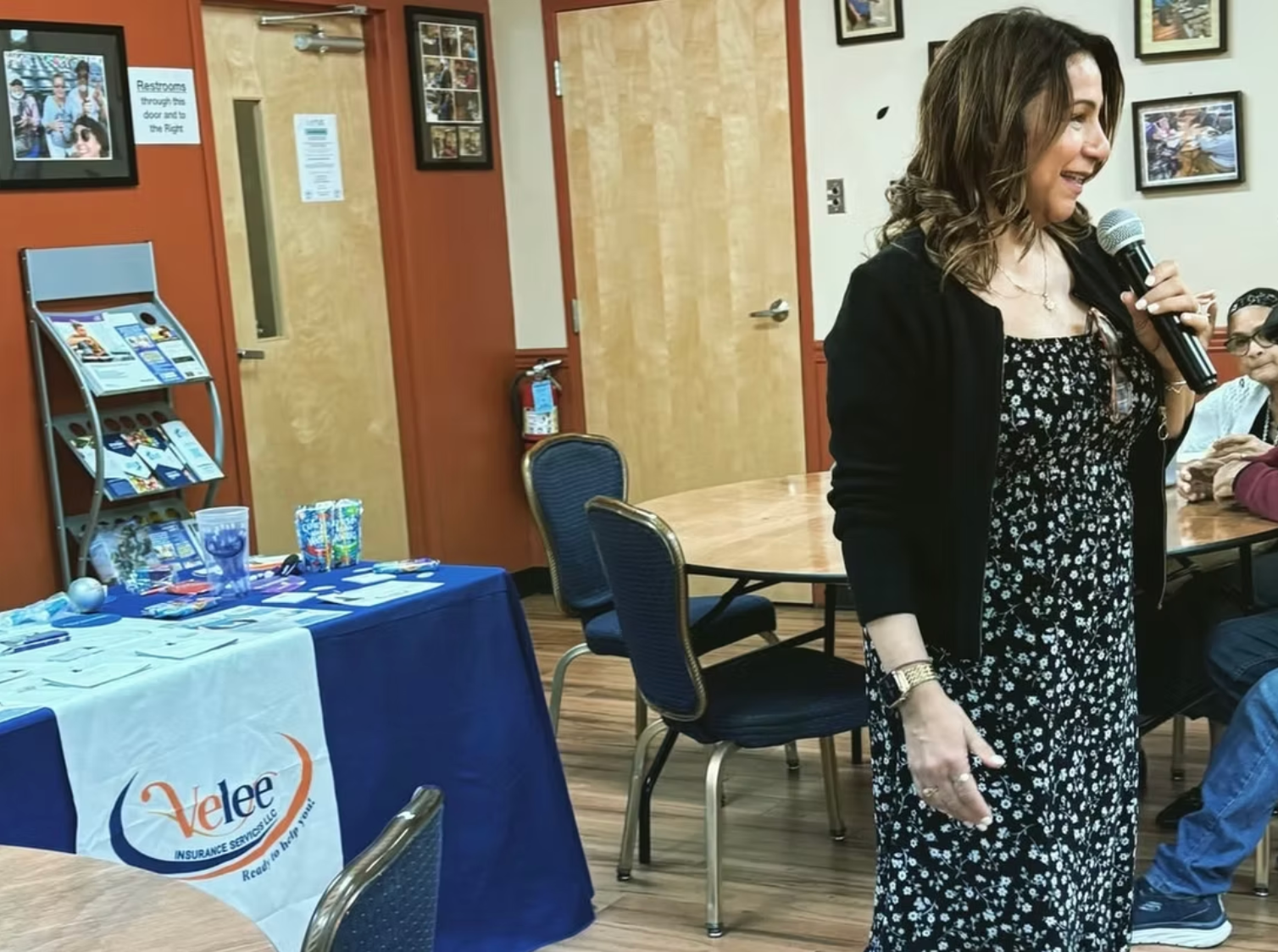A woman in a floral dress and black cardigan speaks into a microphone at an event in an office or community room.