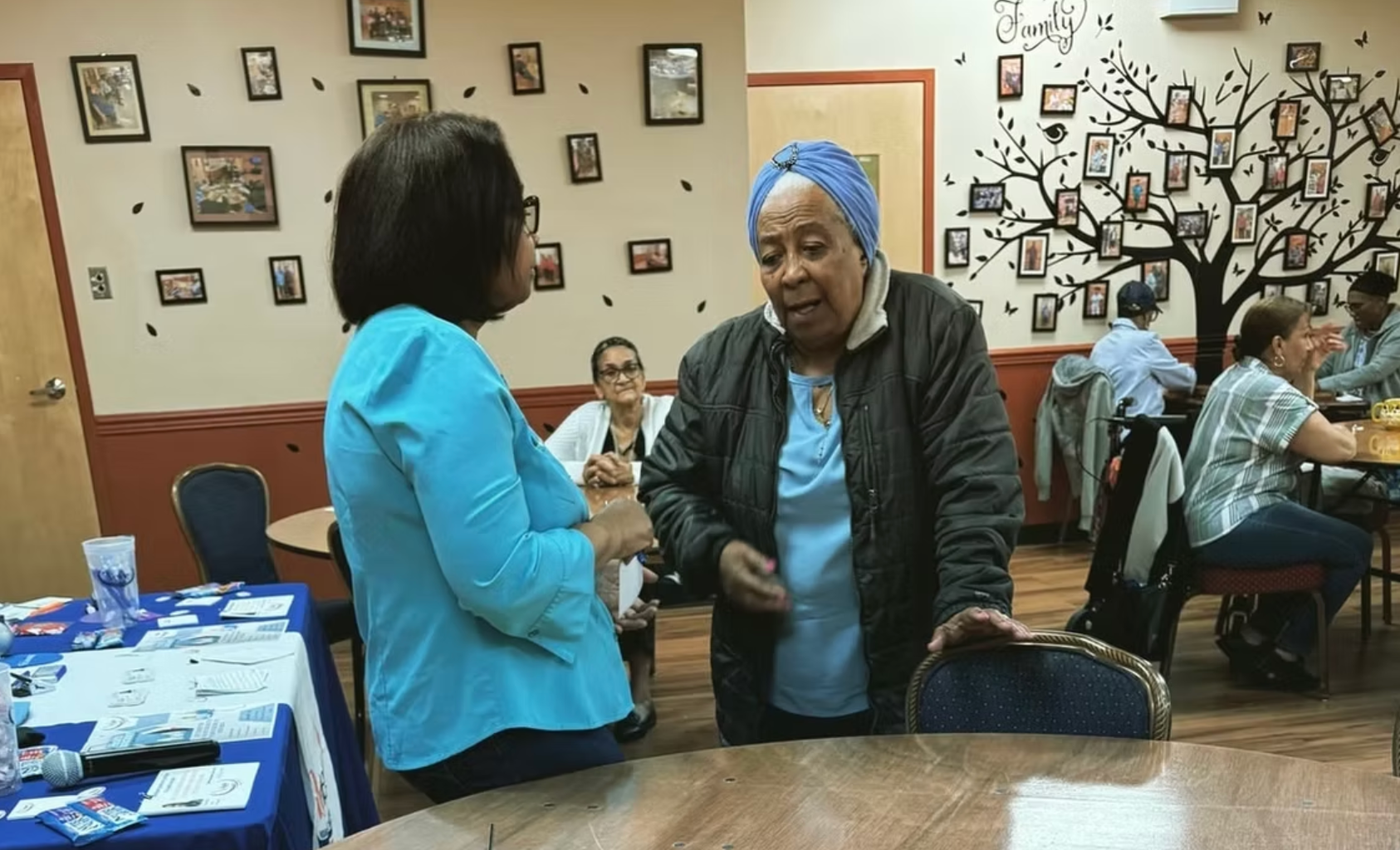 Two people talk in a room with a wall decorated with photos and a mural of a tree. Other people sit at tables nearby.