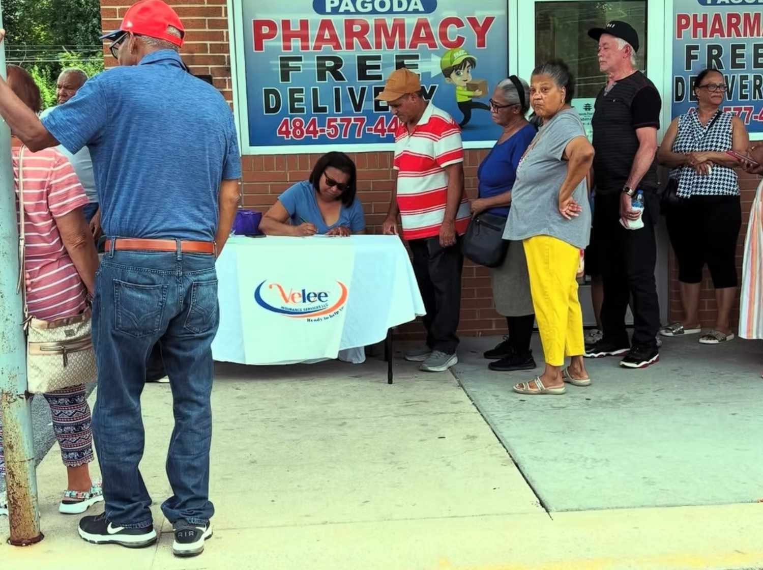 A line of people waits outside Pagoda Pharmacy, where someone is seated at a table filling out forms.
