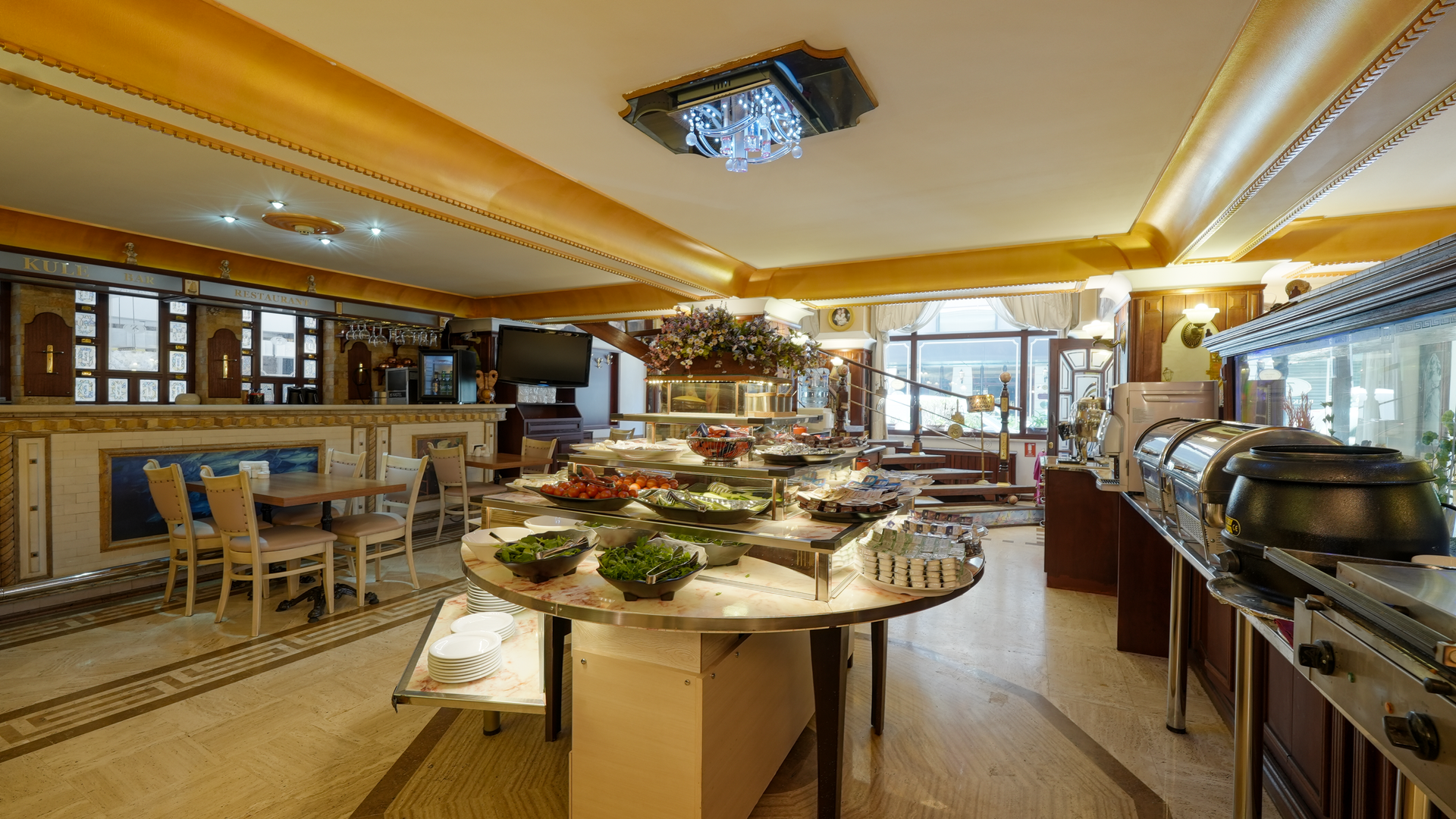 A hotel dining area with a round buffet table in the center, chairs at side tables, and warm, golden lighting.