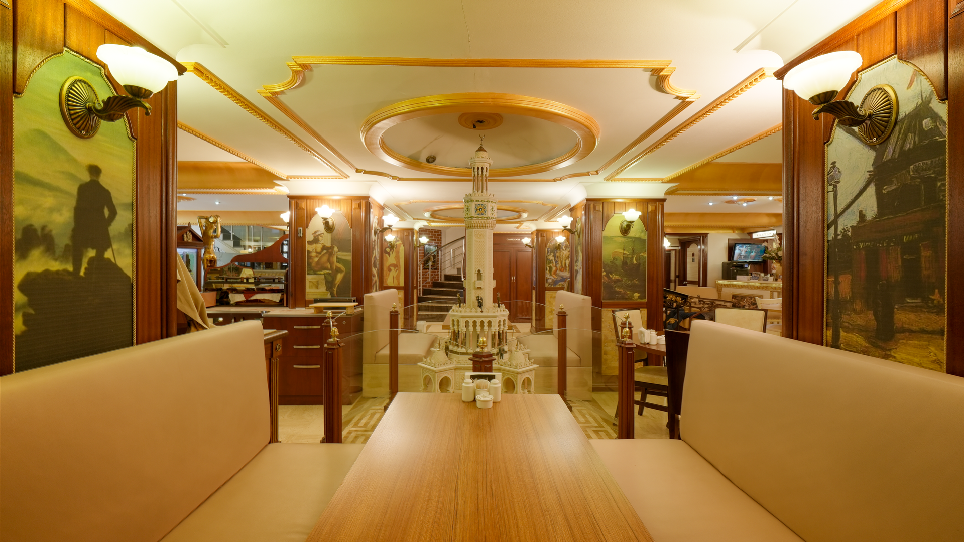 A formal restaurant interior featuring a central wooden dining table with beige booths, ornate gold trim, and wall art.
