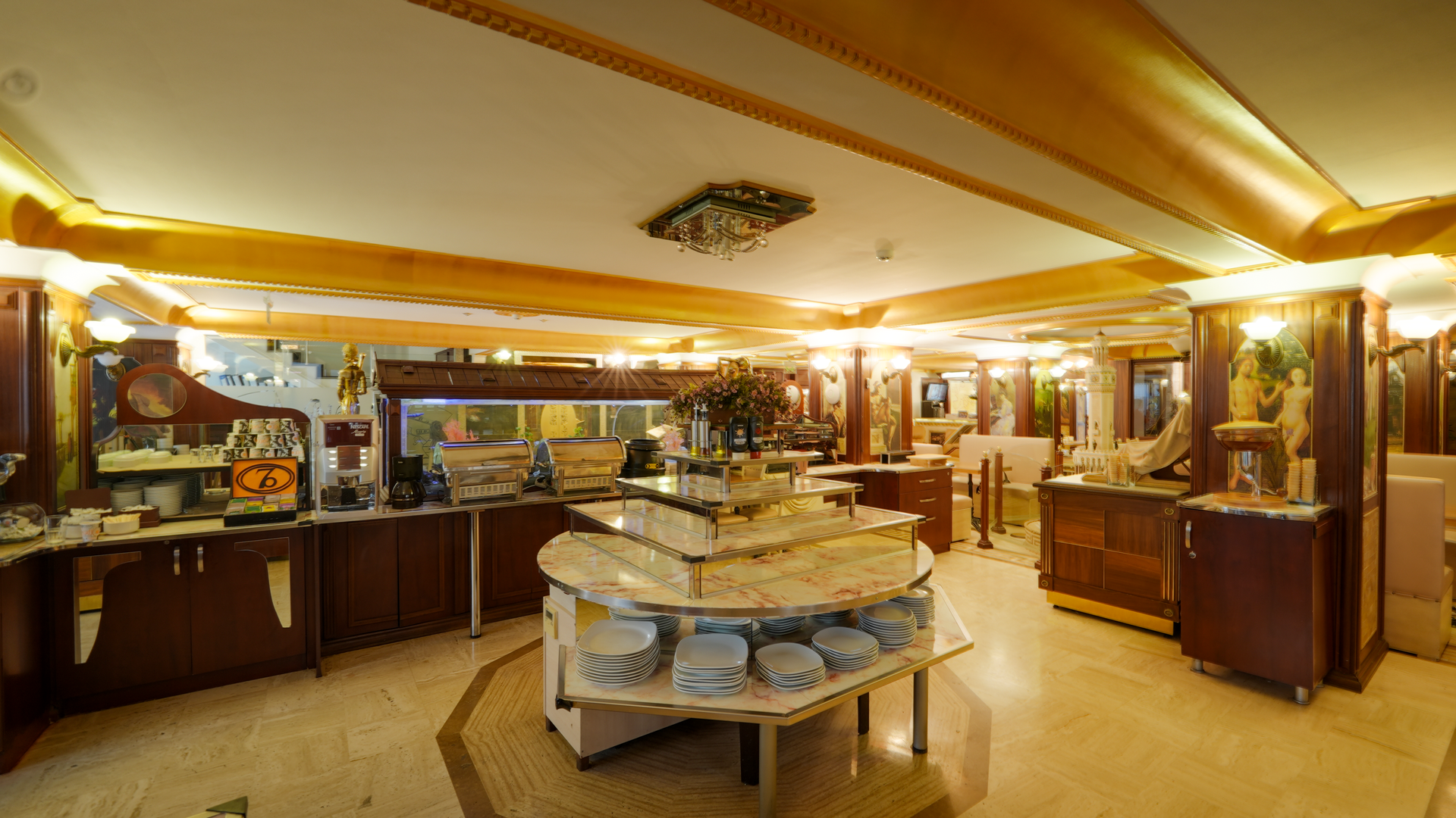 A buffet station in a warm-toned hotel dining area, featuring a tiered display of plates and food items on a central table.