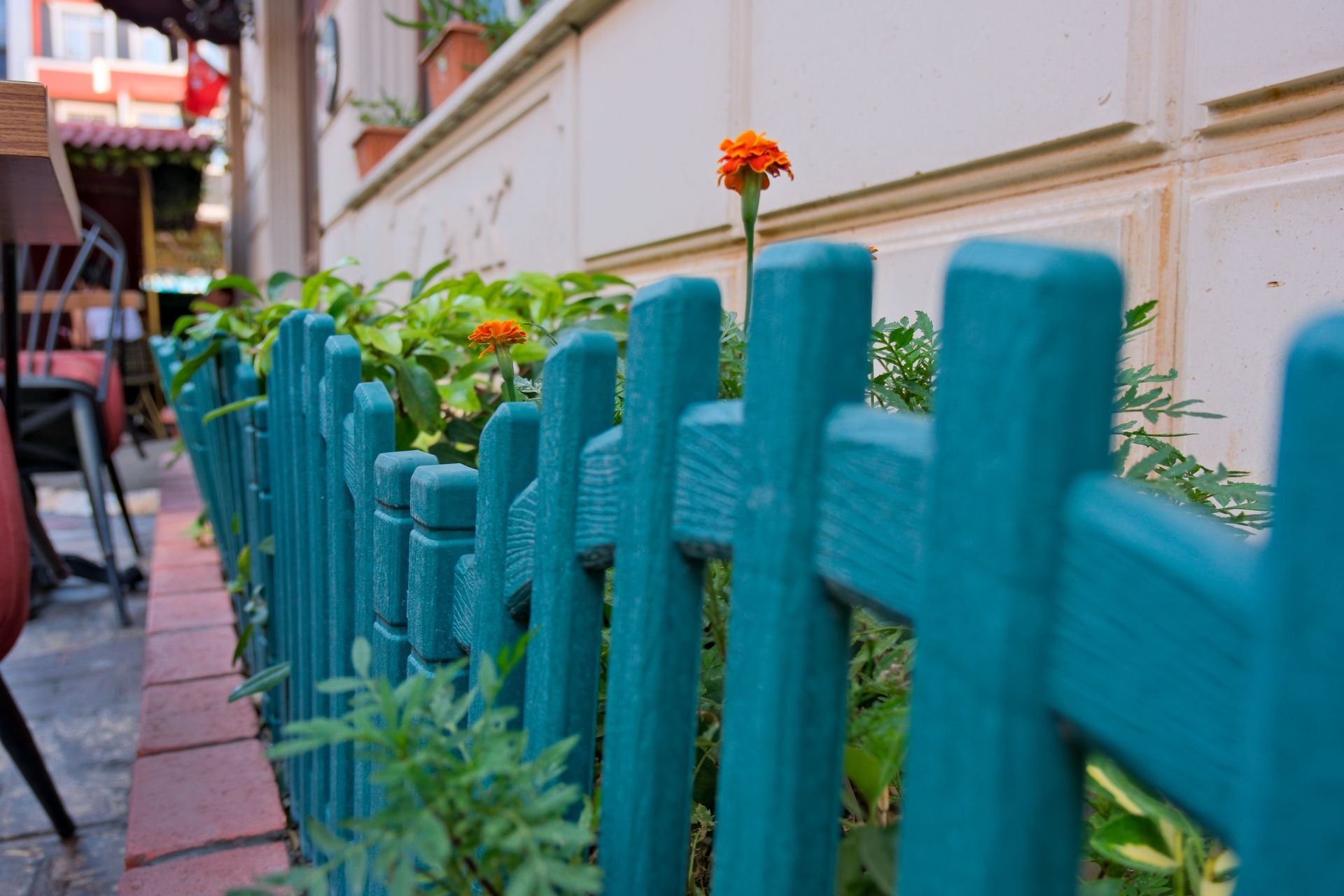 A low, teal wooden fence borders a flower bed with green plants and orange marigolds in front of a building. İzmir Park Hotel