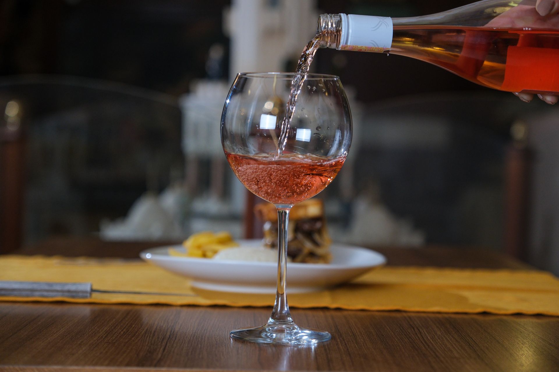 Rose wine being poured from a bottle into a glass on a table with a blurred plate of food in the background.