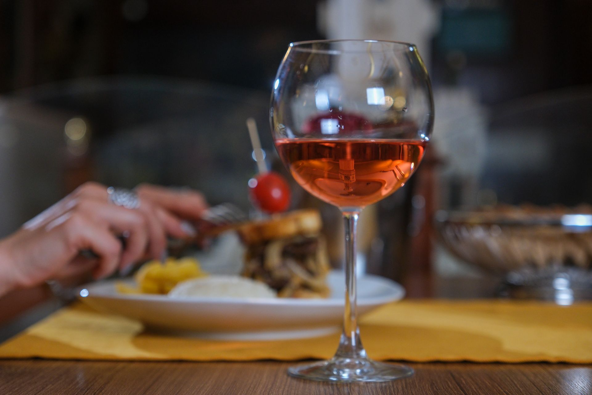 A glass of rose wine sits in the foreground with a blurred plate of food and hands using cutlery in the background.