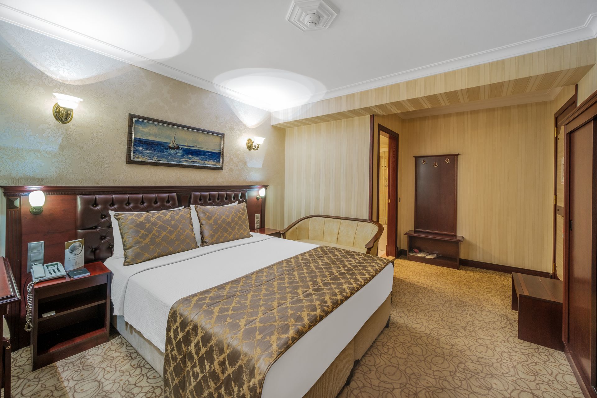 Park Hotel's tidy bedroom with a queen bed, beige patterned carpet, light walls, a sofa, and a framed seascape painting.