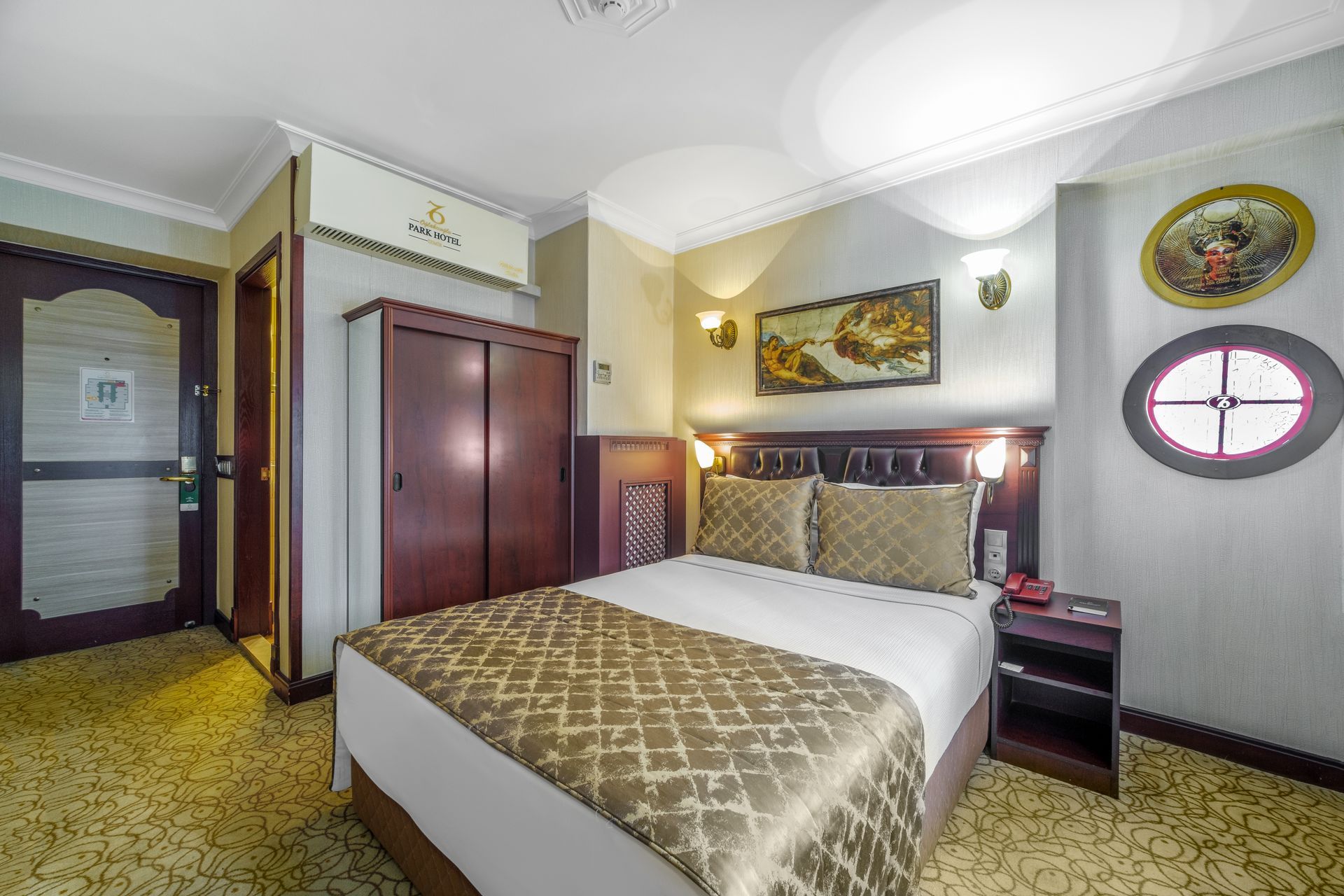 İzmir Park Hotel's room with a queen bed, brown wardrobe, beige carpet, two wall lamps, paintings, and a round window.