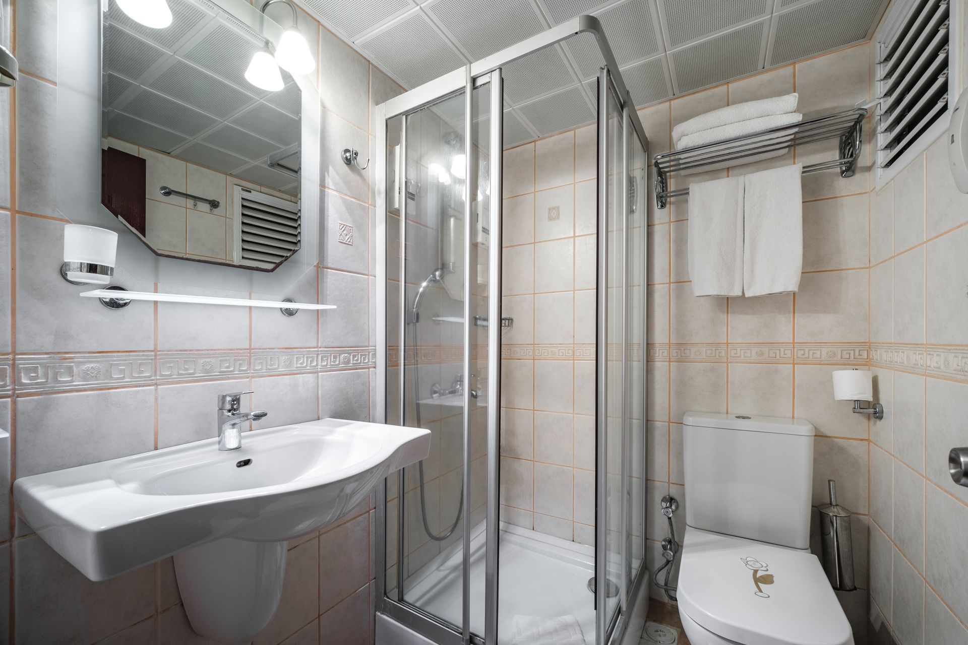 A modern tiled bathroom featuring a pedestal sink with a mirror, a glass-enclosed shower, and a toilet with hanging towels.
