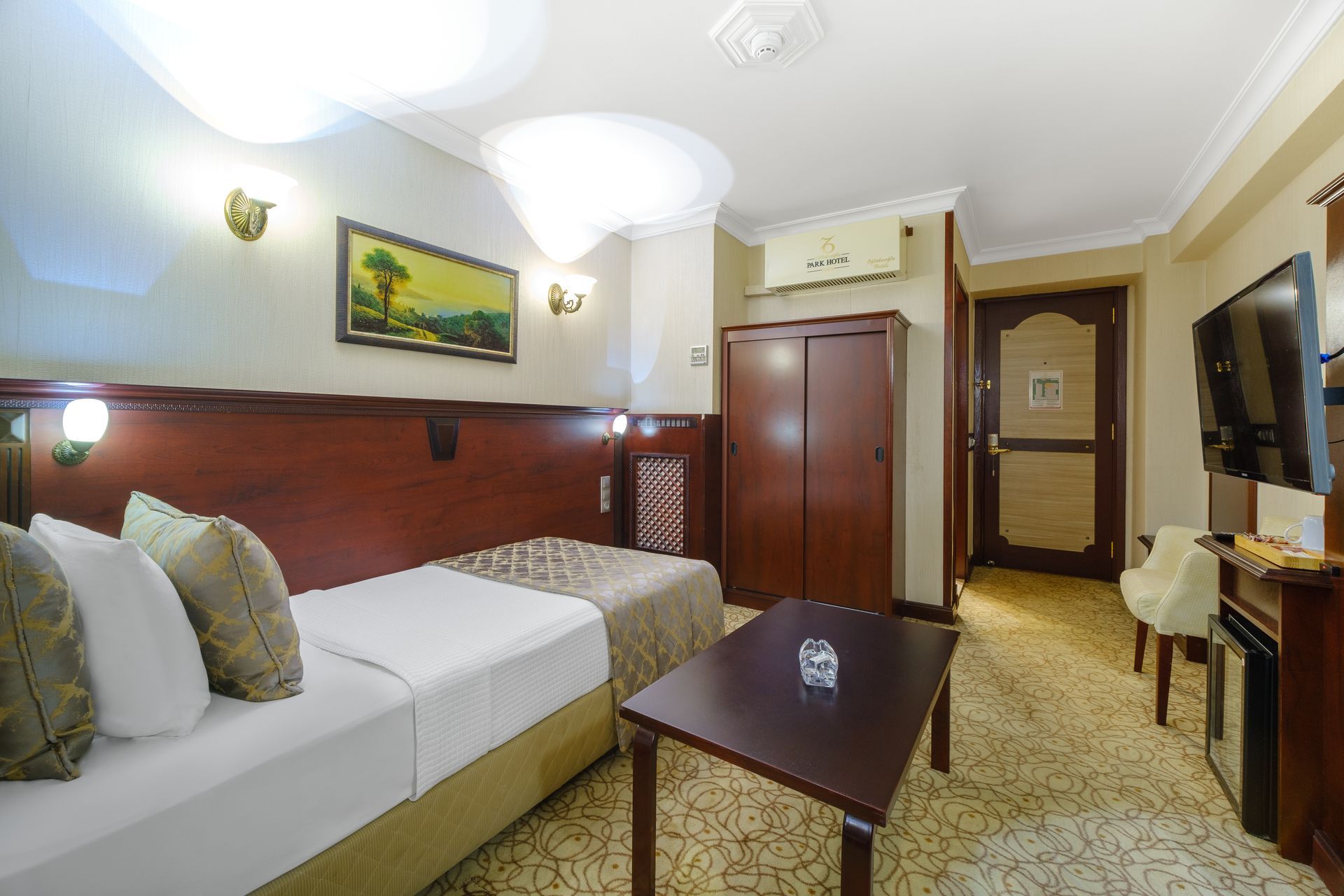 İzmir Park Hotel's room with a single bed, a wooden wardrobe, a coffee table, a flat-screen TV, and patterned beige carpet.