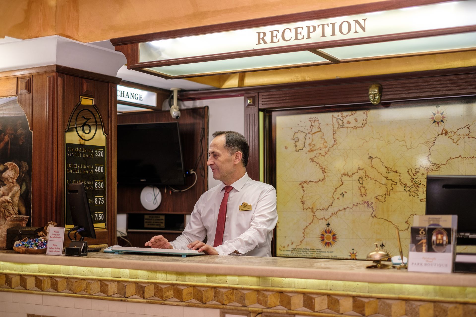 Oğlakçıoğlu İzmir Park Hotel's receptionist in a light shirt and red tie works at a desk in front of a wall-mounted map.