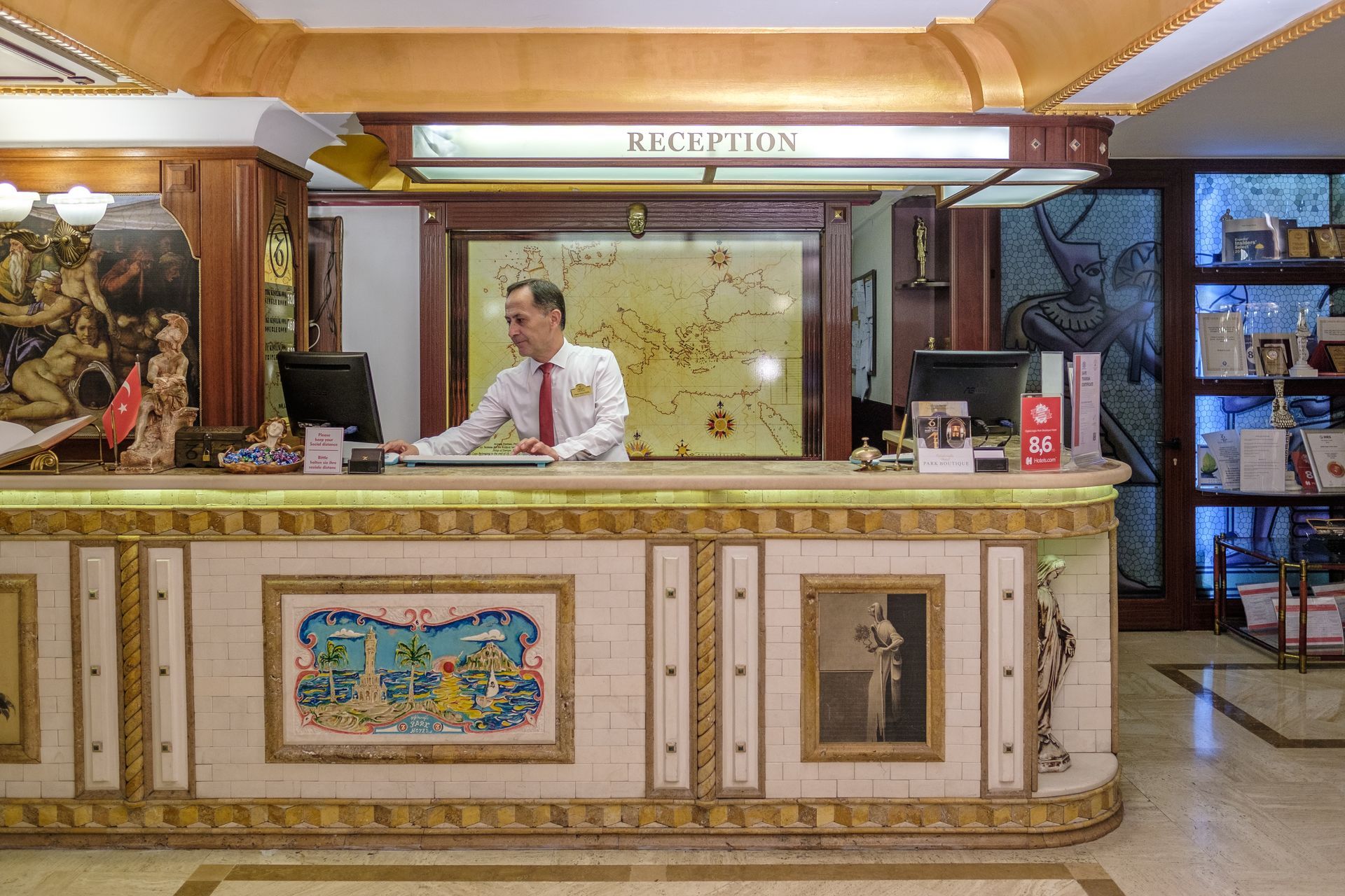 İzmir Park Hotel's receptionist in a white shirt and red tie stands behind an ornate reception desk with tiled artwork.