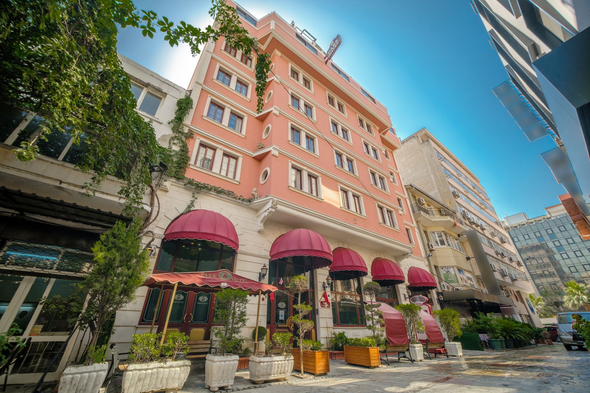 Oğlakçıoğlu İzmir Park Hotel hotel with red awnings and decorative ivy, situated on a sunny city street.